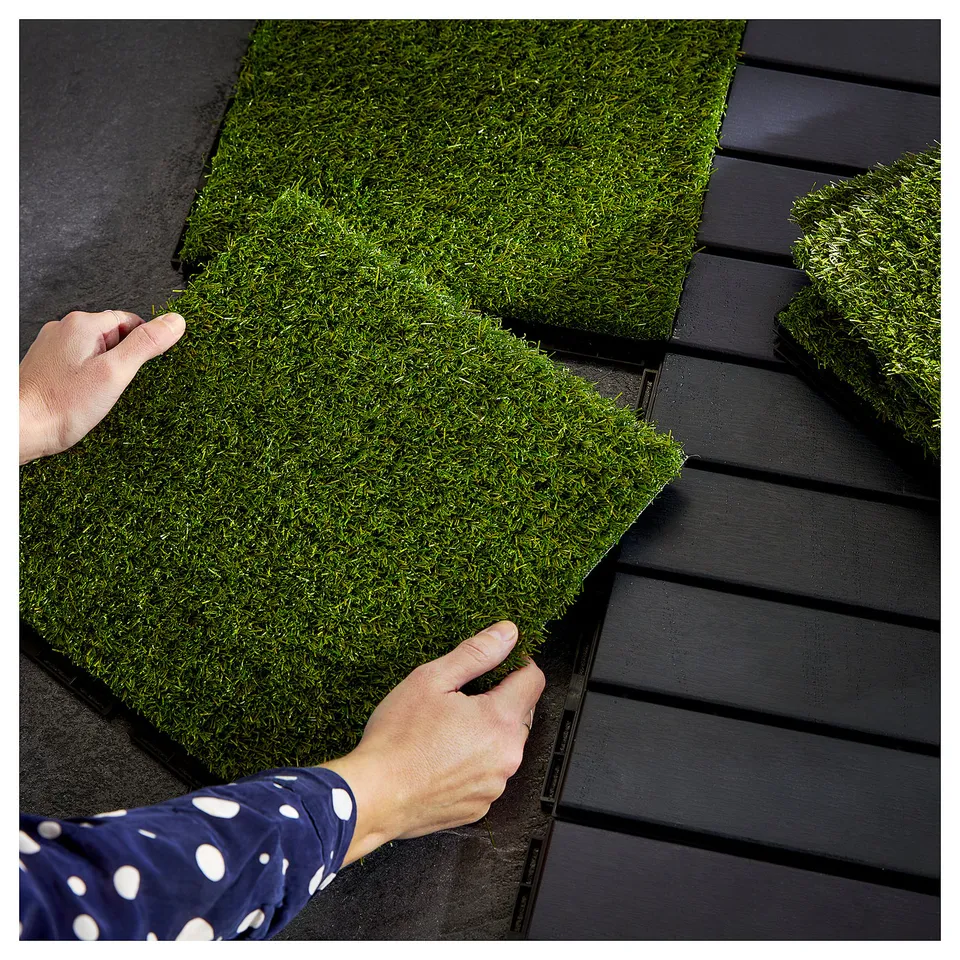 Person placing green artificial grass panels on dark floor.