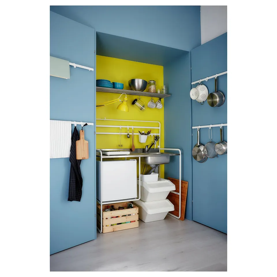 Modern blue and yellow kitchenette with stainless sink, white storage, hanging pots, and wooden crate.