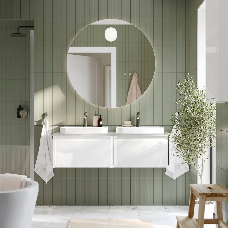 Modern bathroom with round backlit mirror, green tiled walls. White vanity, towels, soft lighting. Plant and bathtub visible.