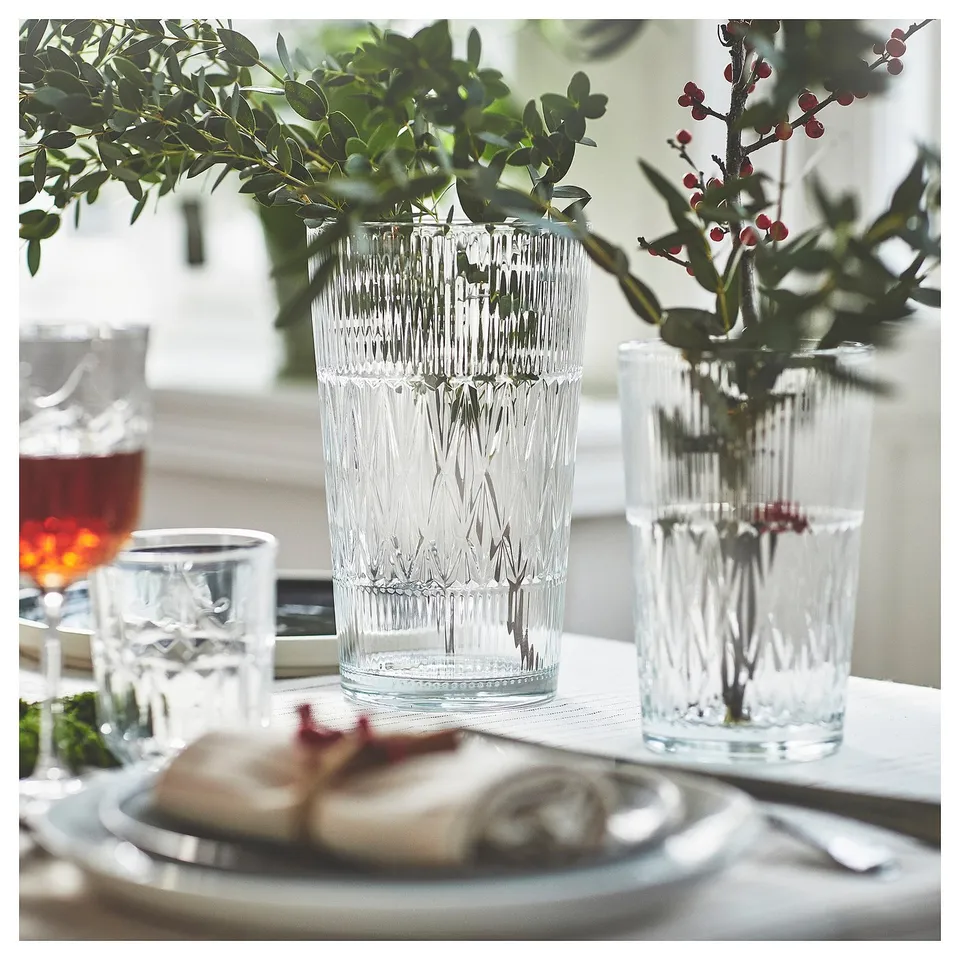 Three elegant, clear glass vases with diamond patterns hold greenery and berries, set on a white table with a plate of food.