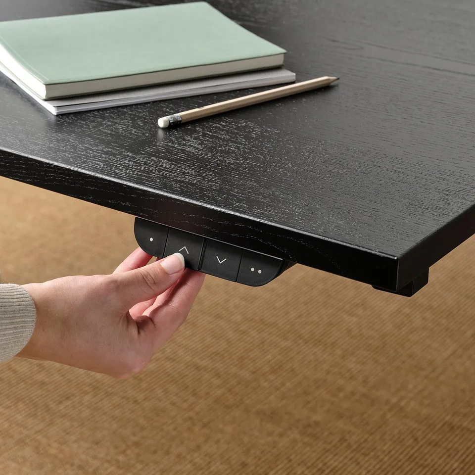 Person adjusting height of black sit-stand desk using hand control. Green notebook and pen on top.