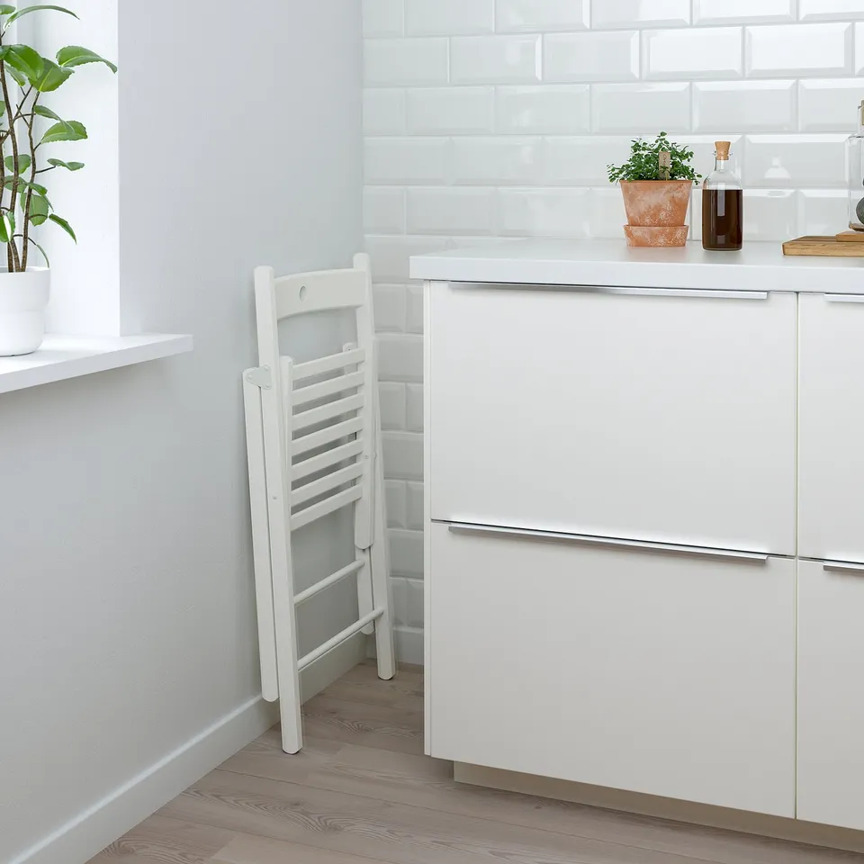 White kitchen setup with folded chair and potted plant on shelf.