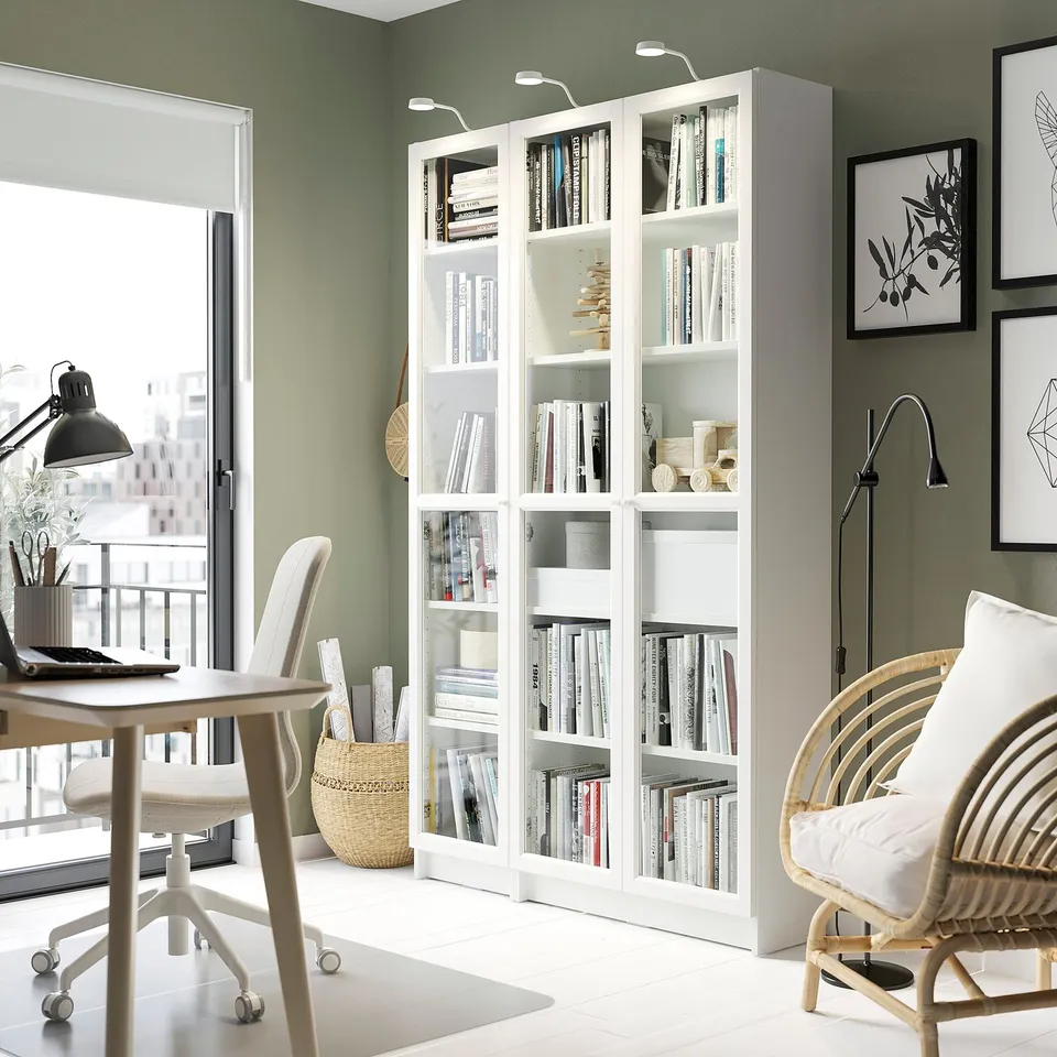 White BILLY bookshelf with glass doors in home office. Wooden & wicker chairs nearby. Green wall with art.