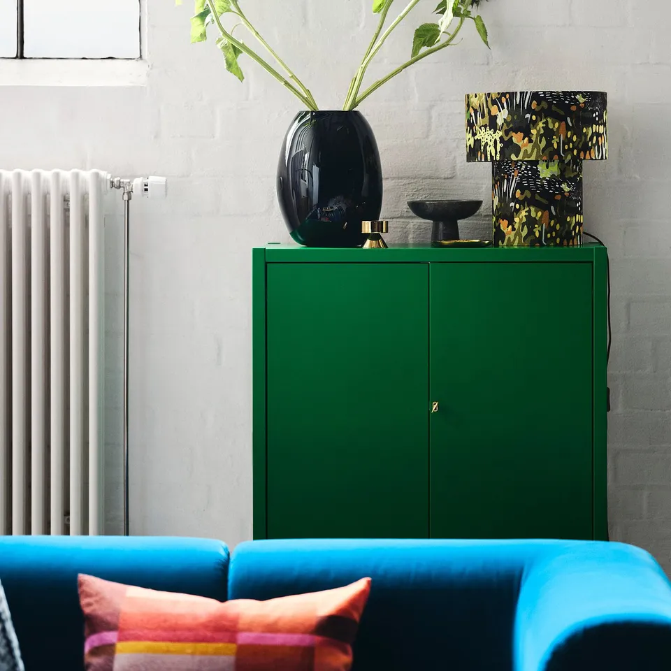 Bright blue sofa with orange pillows in front of green STOCKHOLM 2025 cabinet; black vase, colourful lamp, and plant on top.