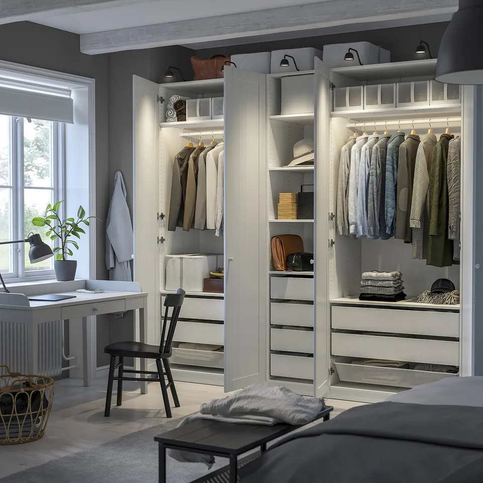 Well-organized bedroom with an open PAX wardrobe showcasing clothes, accessories, and lighting. A desk, chair, and plant are also visible.