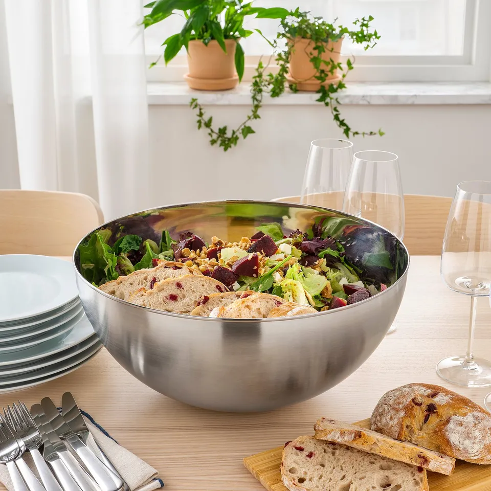 Large stainless-steel salad bowl on a dining table surrounded by glasses, plates, cutlery, and bread, ready for serving.