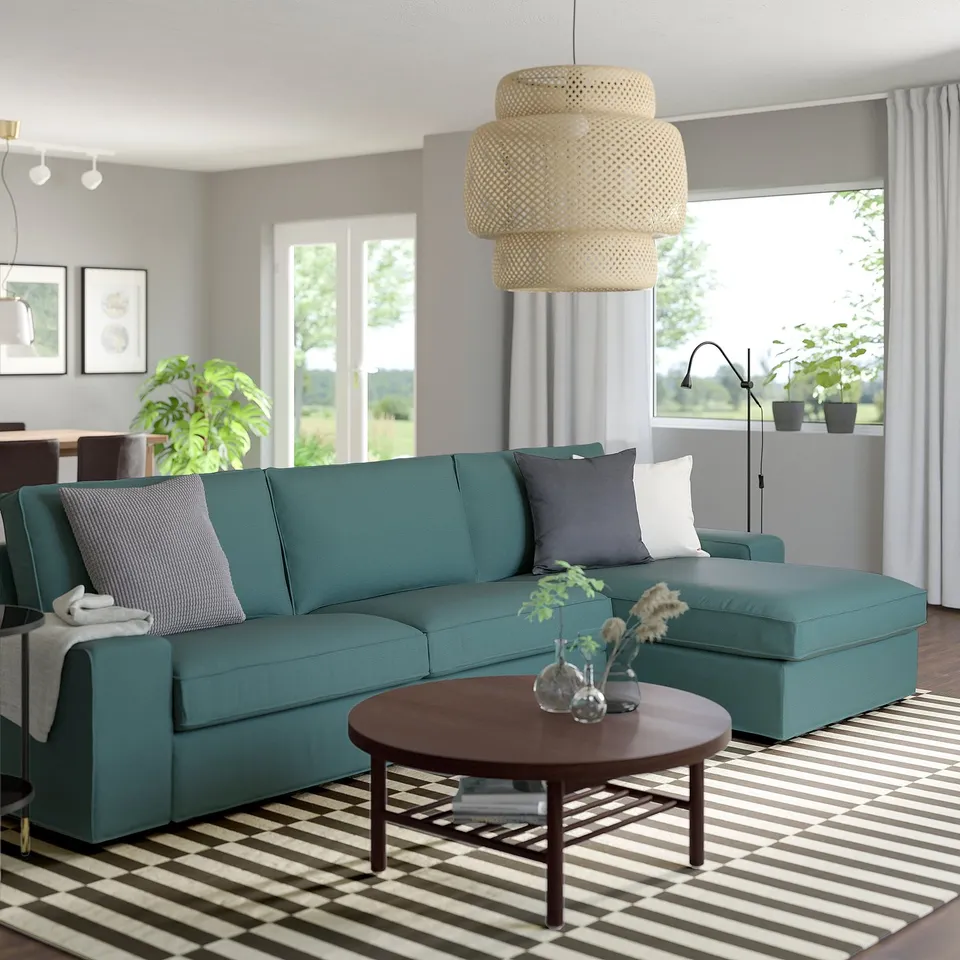 Modern living room with large teal KIVIK sofa, wooden coffee table, striped rug, hanging light, plants, and large windows.