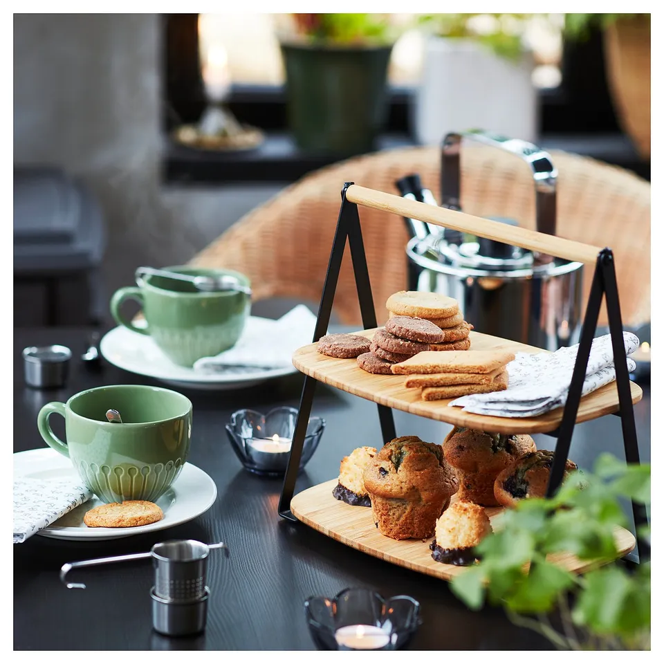Two-tier bamboo serving tray with black metal stand holds muffins and cookies. Green cups and small bowls surround on dark table.