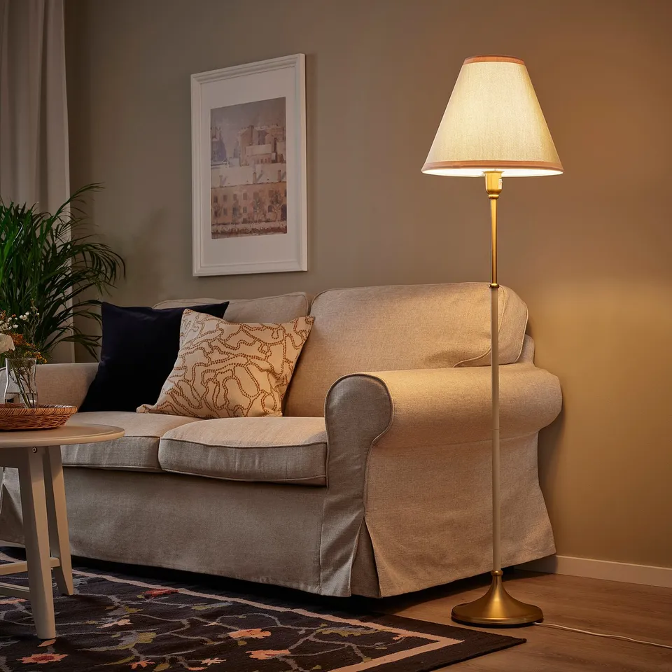 A cosy living room with a beige sofa, warmly lit by a golden FLYGHÖJD floor lamp, standing tall and elegant beside it.