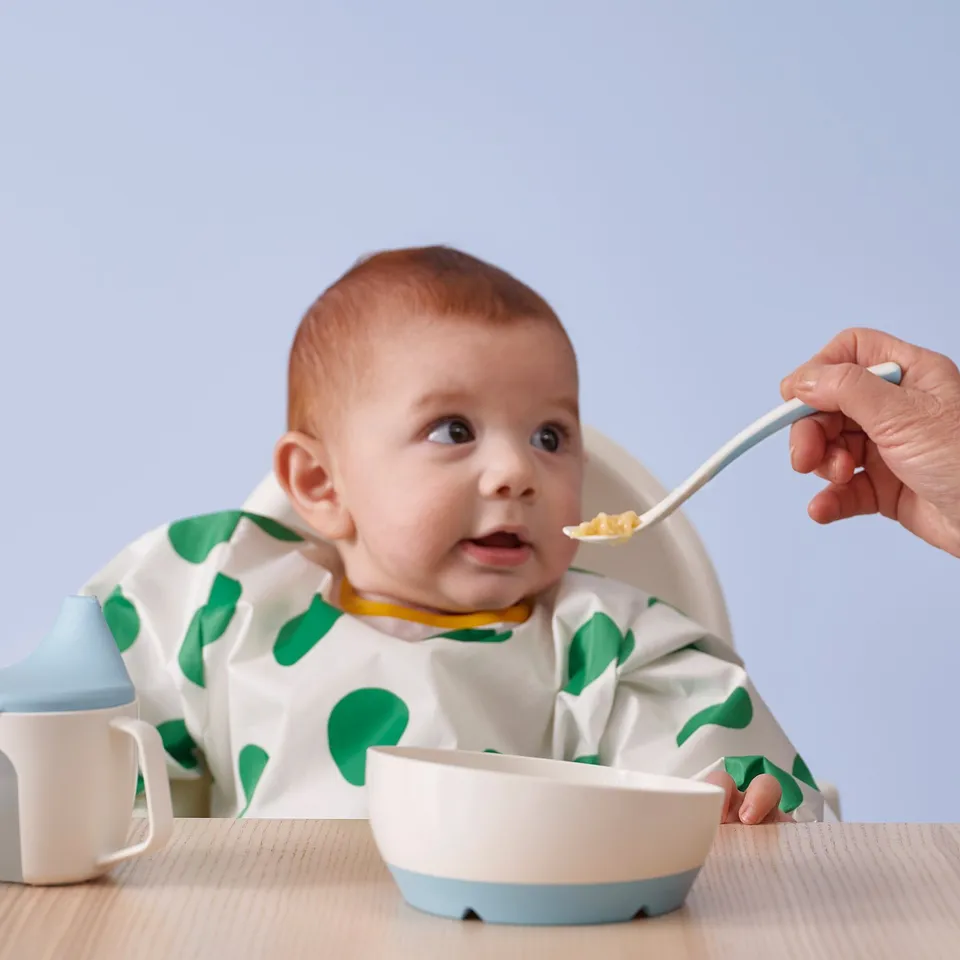 Baby in high chair, wearing bib, being fed from a blue bowl using a spoon. Bowl has wide base, stays stable on the table.