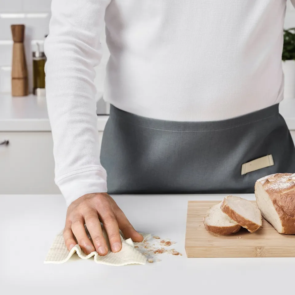 Person wiping counter with TRIXIG cloth, cleaning up crumbs beside a loaf of bread on a cutting board.