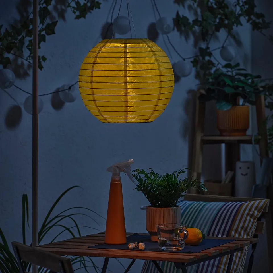 Outdoor cosy scene with SOLVINDEN lantern above table with plant, spray bottle, glass, and orange.