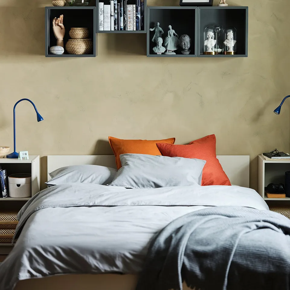 Modern bedroom with grey bedding, orange pillows, blue wall shelves holding books, plants, and statues.