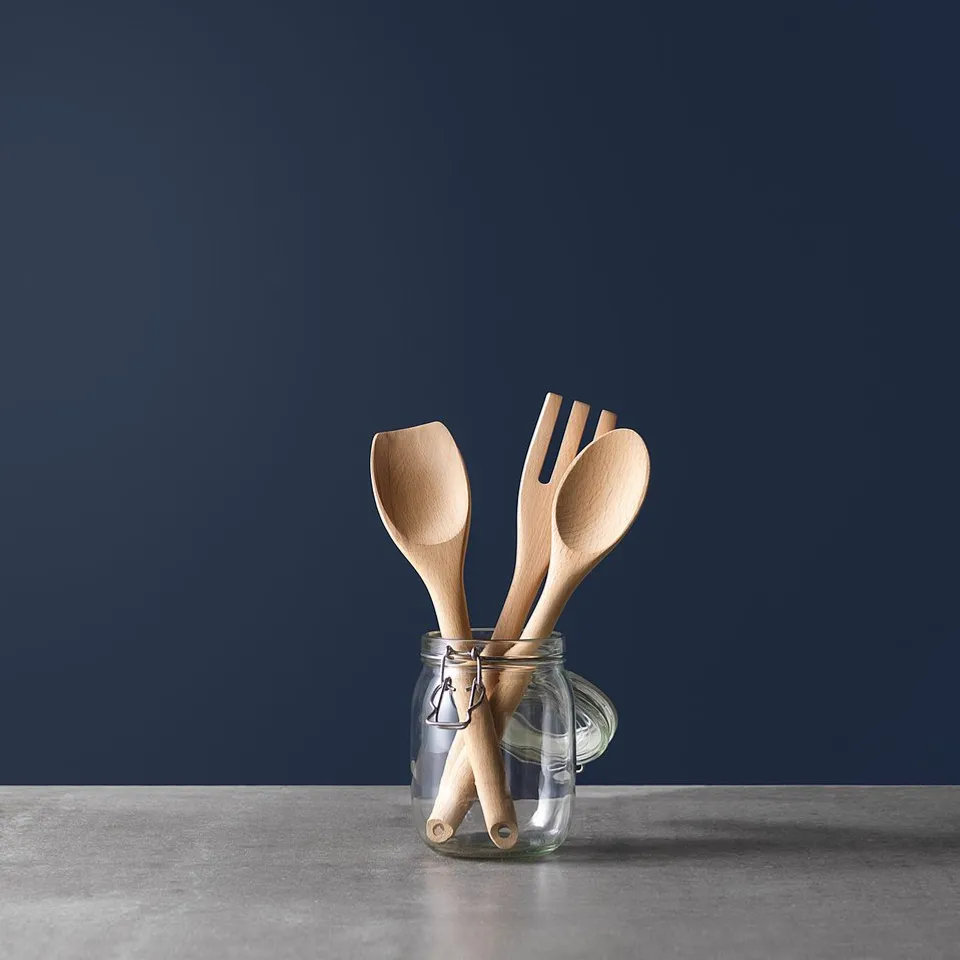 A jar holds wooden RÖRT utensils: spoon, spatula, fork. Dark blue background, natural wood colour, smooth and simple design.