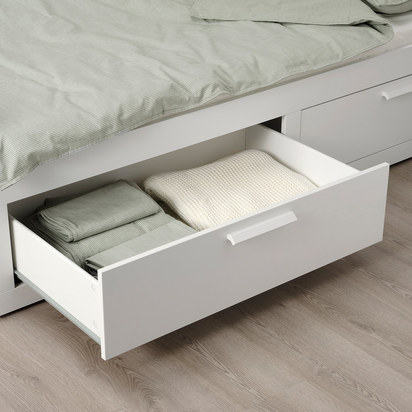 A white under-bed drawer open, storing folded blankets on a wooden floor, providing space-efficient storage.
