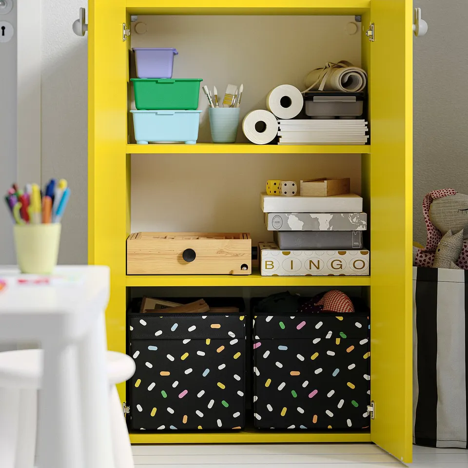 Yellow storage cabinet with multiple shelves holding office supplies, games, and storage bins. Adjustable feet for floor levelling.