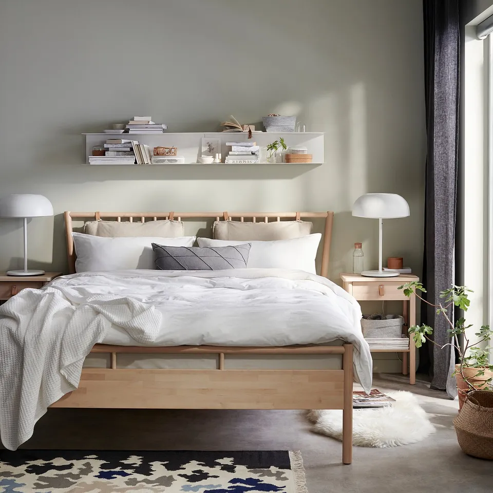 Modern bedroom with wooden bed, white linens, grey pillows, bookshelves, lamps, and patterned rug.