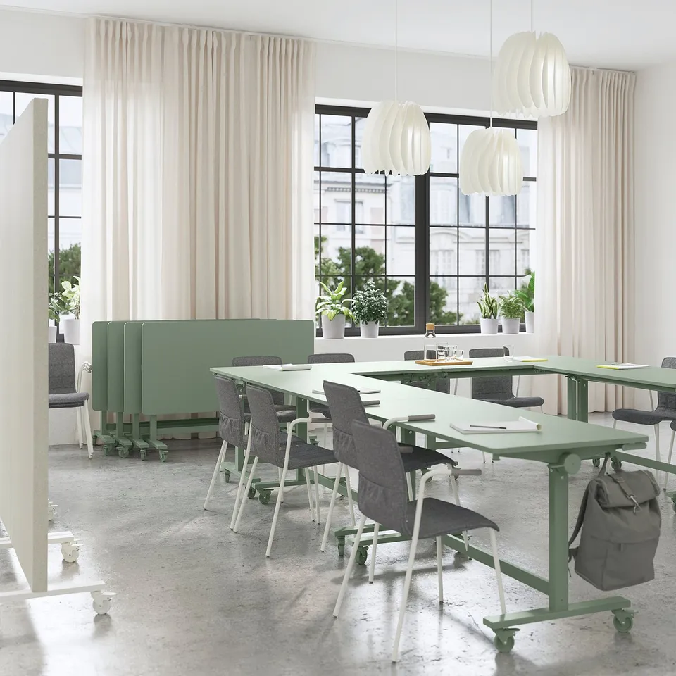 Modern office with light green acoustic panels and tables. Chairs, bags, and plants. Rectangular tables with hanging lights, large windows.