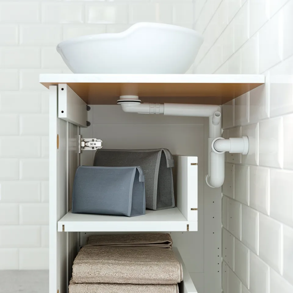 White cabinet under sink, holding grey baskets and folded towels, showing pipe access.