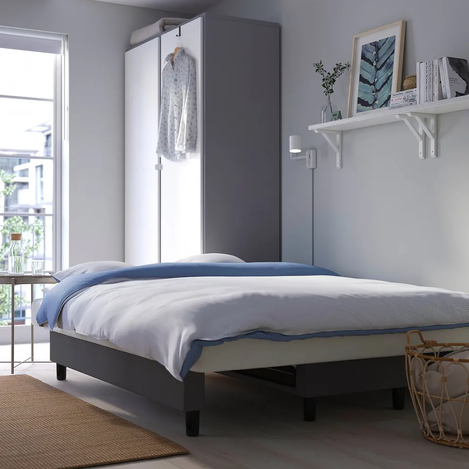 Bed folded out from sofa, showcasing practical storage beneath seat. White and blue bedding, wall shelf with decor, and natural light through window.