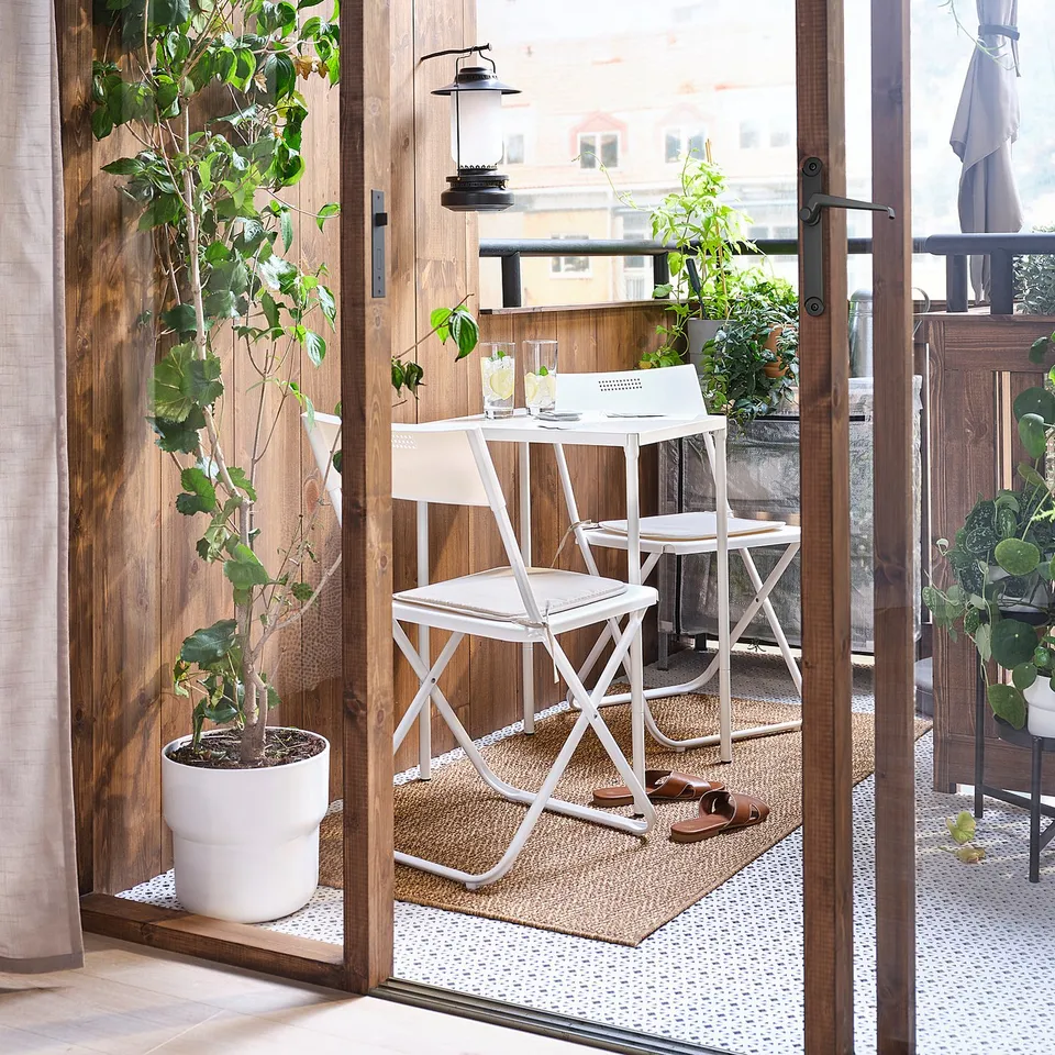 Small outdoor setup with white FEJAN table and chairs. Wooden deck, green plants, and sliding doors. Minimalistic and modern design.