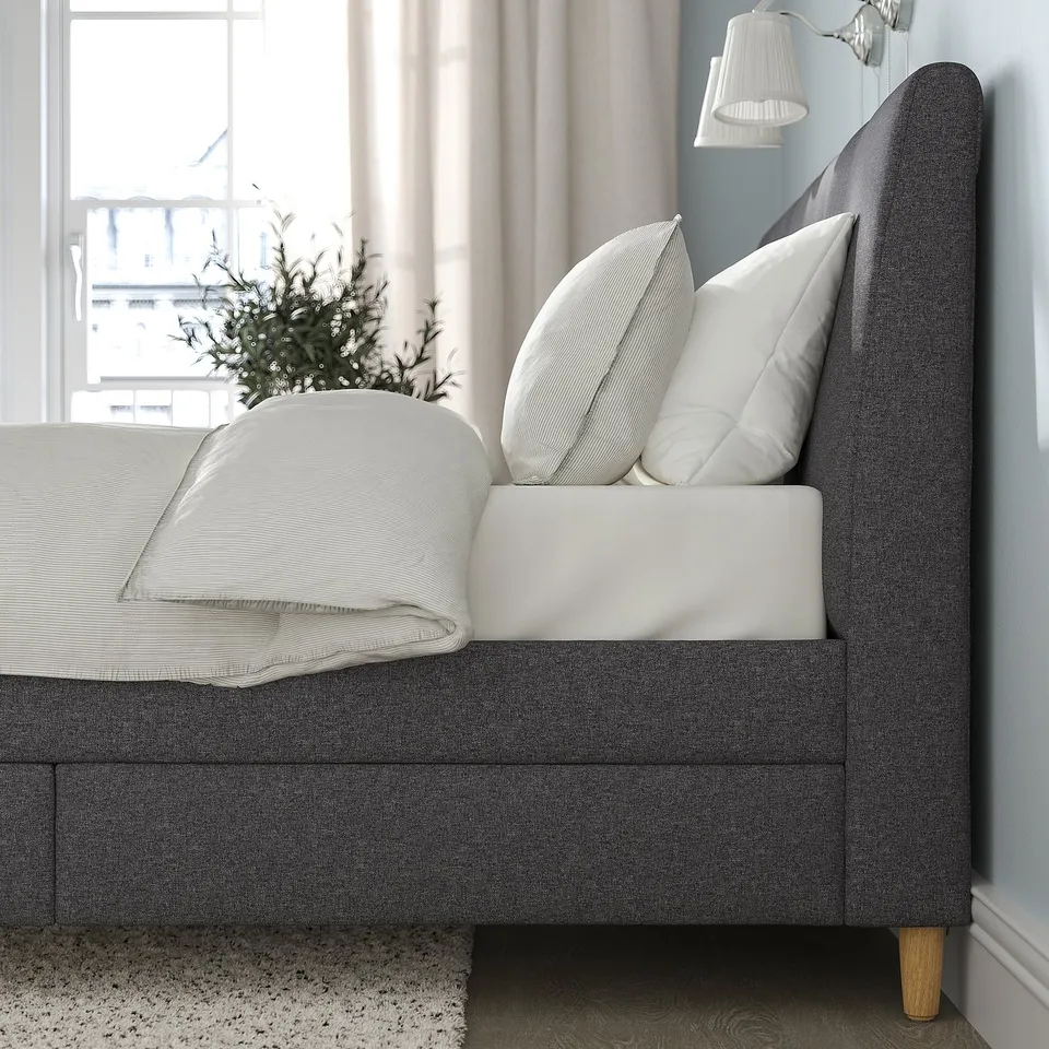 Bed with slanted headboard, upholstered in grey. Drawer storage under bed, matching frame legs.