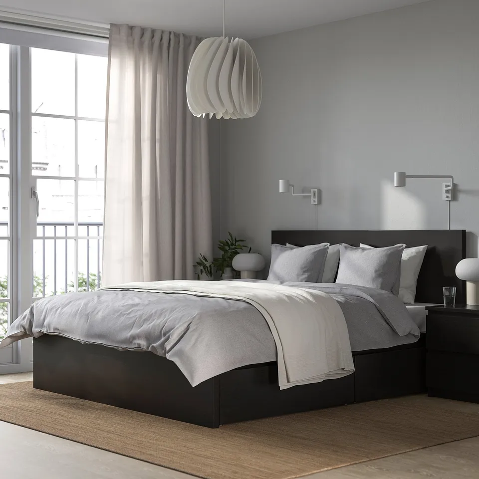 Minimalist bedroom: black bed, white linens, grey curtains, unique light. Light blue walls, storage, plant, large window.