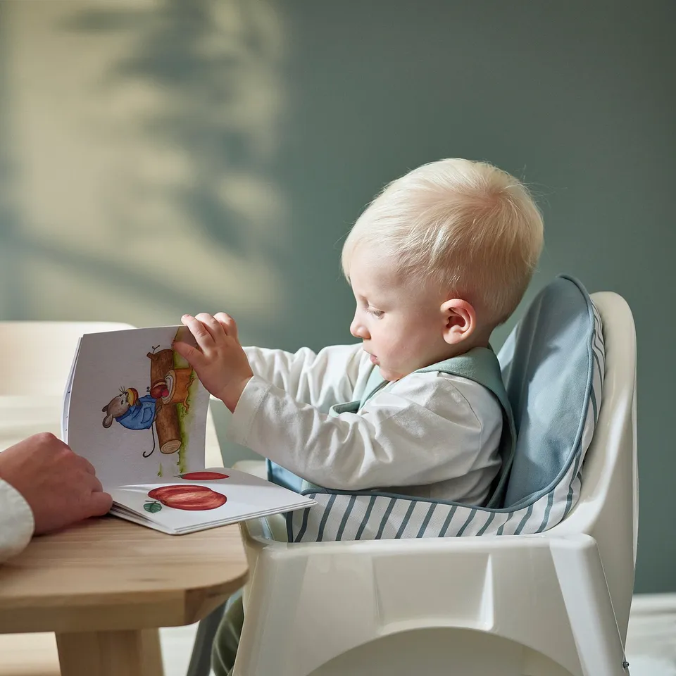 Baby sitting in a high chair, reading a book with support from an inflatable cushion.