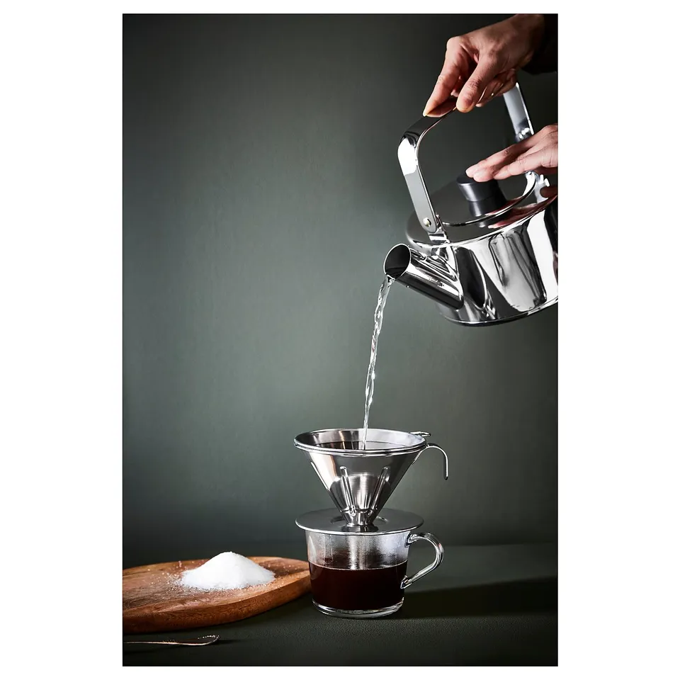 Silver stainless steel coffee dripper pouring into glass mug on table.