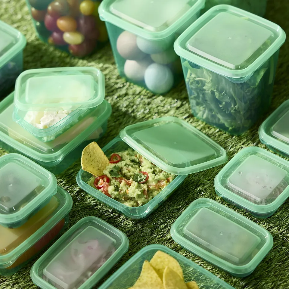 Green plastic food containers with lids, storing various foods. One has guacamole and a chip.