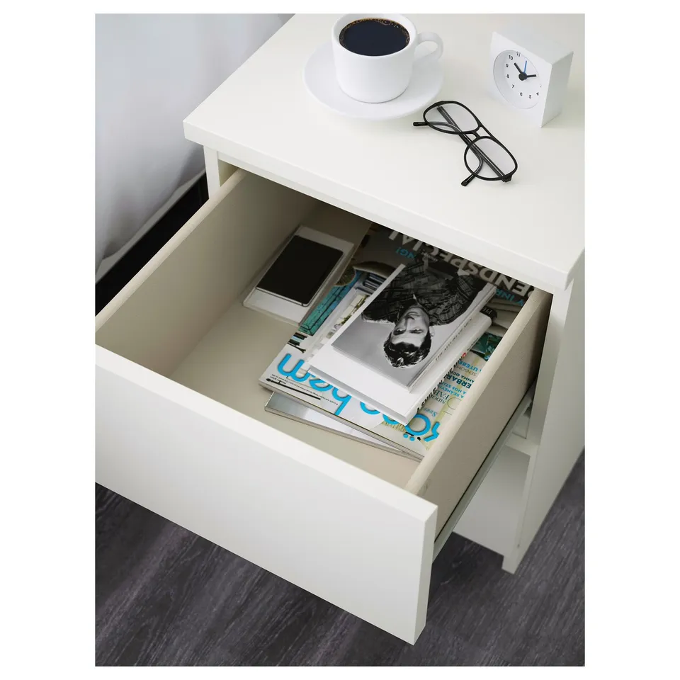 White bedside table drawer open revealing magazines and a smartphone. Top has a cup, glasses, and clock.