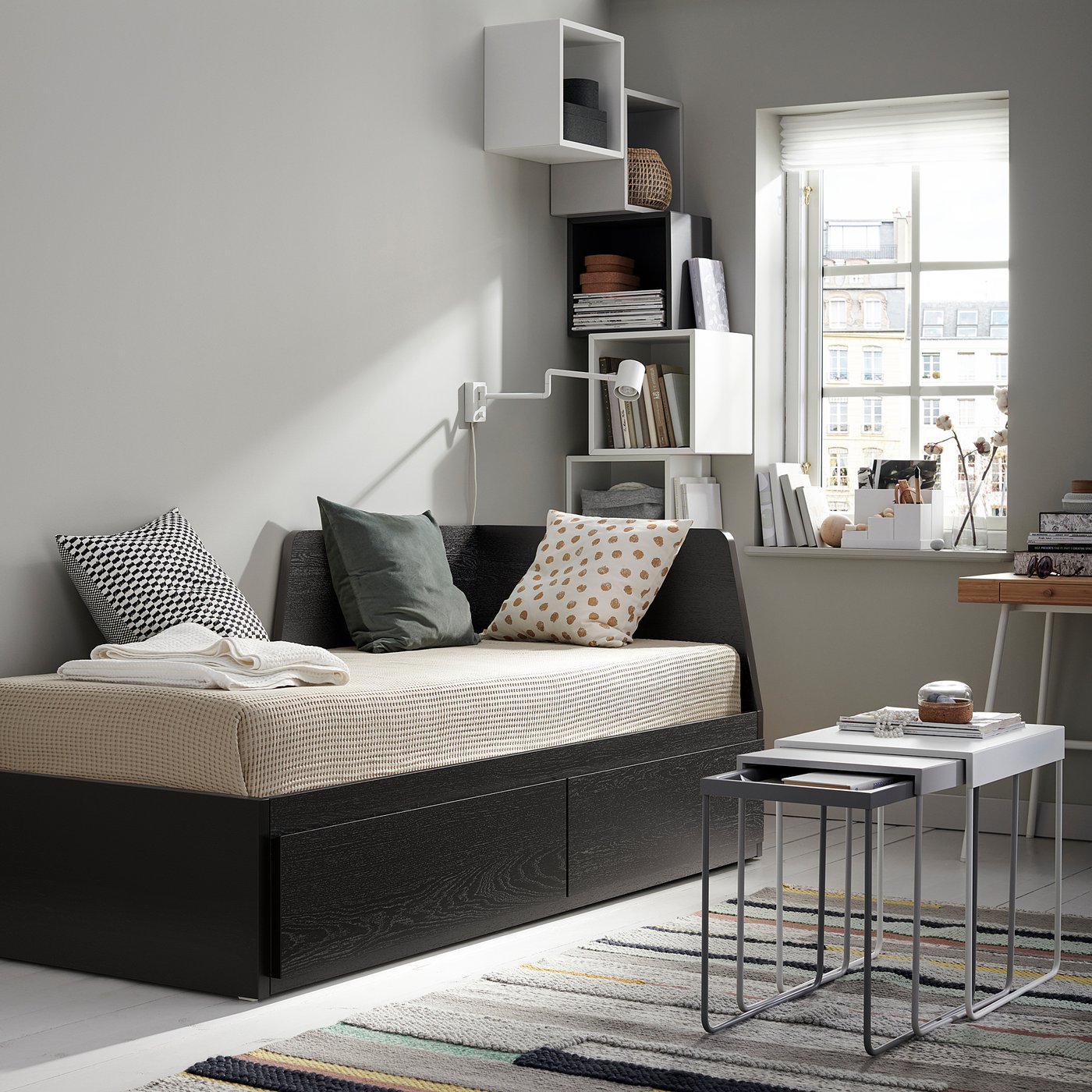Modern living room with a FLEKKE day-bed, white shelves, colourful rug, and metal nesting tables.