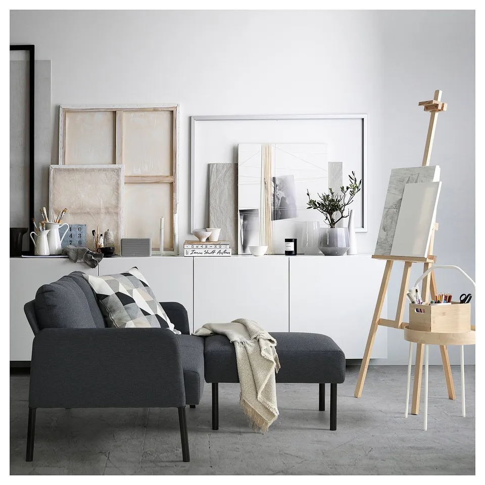 Modern living room with GLOSTAD dark grey sofa and footrest, white sideboard with decorative items, and wooden easel with art supplies.
