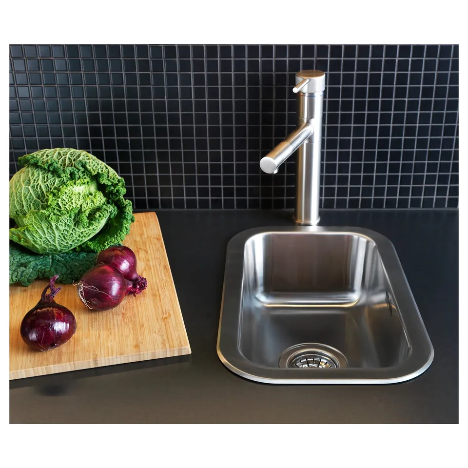 Modern kitchen sink in stainless steel with sleek tap, set against dark tiled backdrop. Vegetables placed on wooden board beside it.