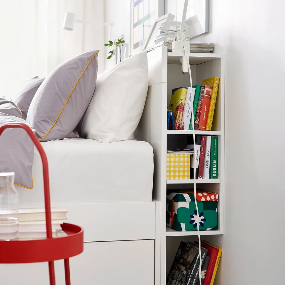 Neat bedroom with white BRIMNES bed, grey pillows, red side table, and shelves filled with colourful books and decorated with a green leafy plant.