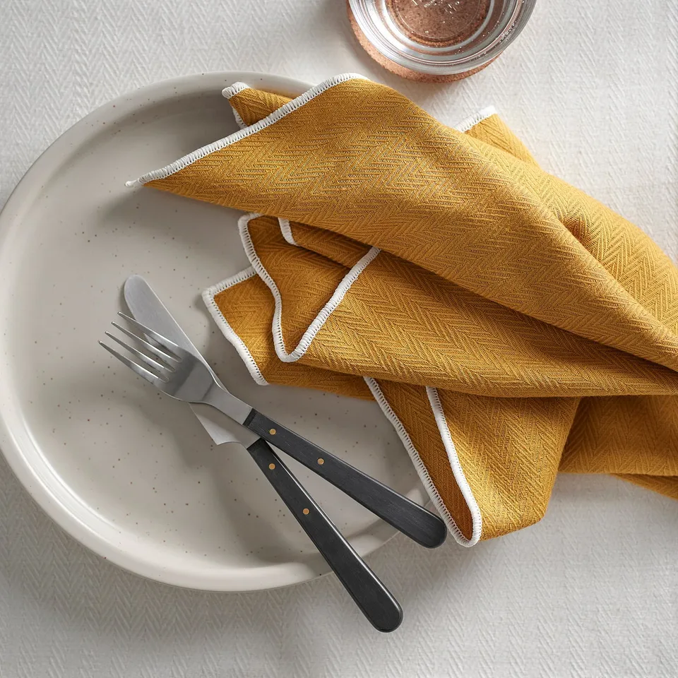 SANDABBORRE mustard napkin with white trim, folded on plate with black-handled fork and knife.