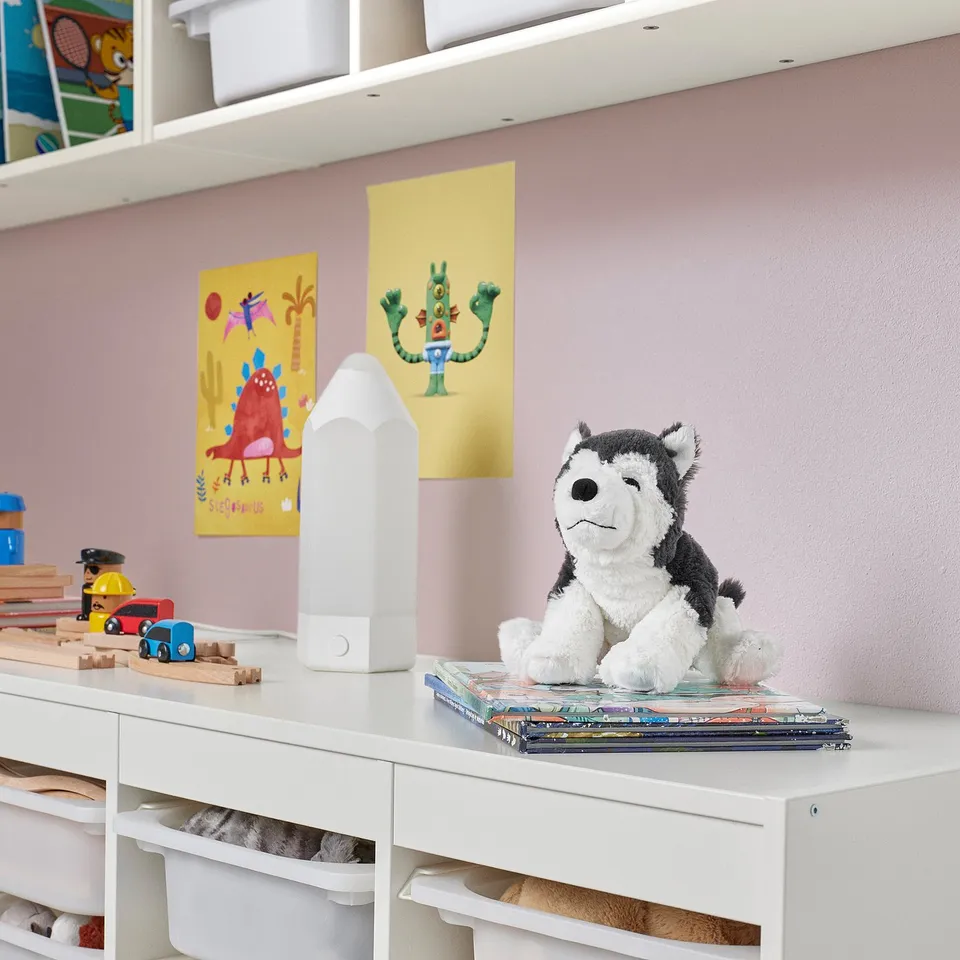Childrens room with white shelves. Colourful artwork, toy cars, blocks, and a stuffed grey and white husky sit atop books, smaller than a lamp.