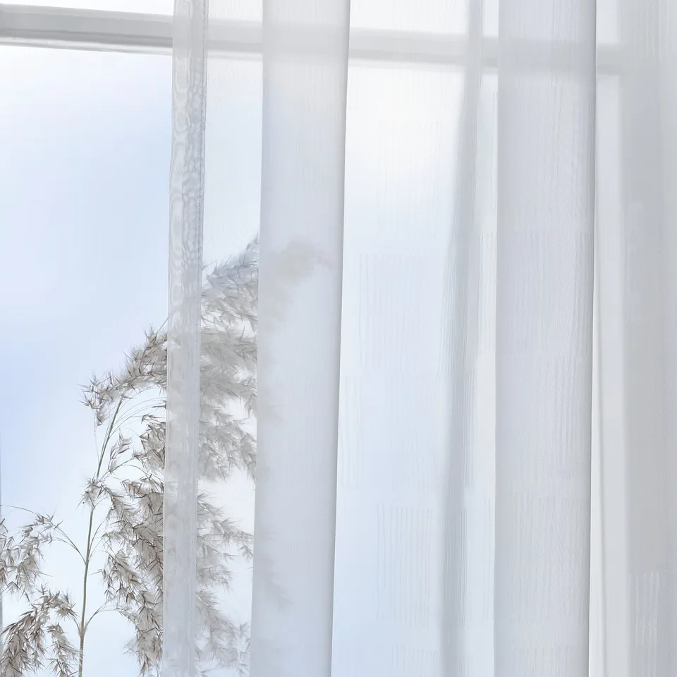 Sheer white curtains with subtle pattern softly diffuse sunlight, creating calm and privacy. Heading tape allows easy pleating and customization.
