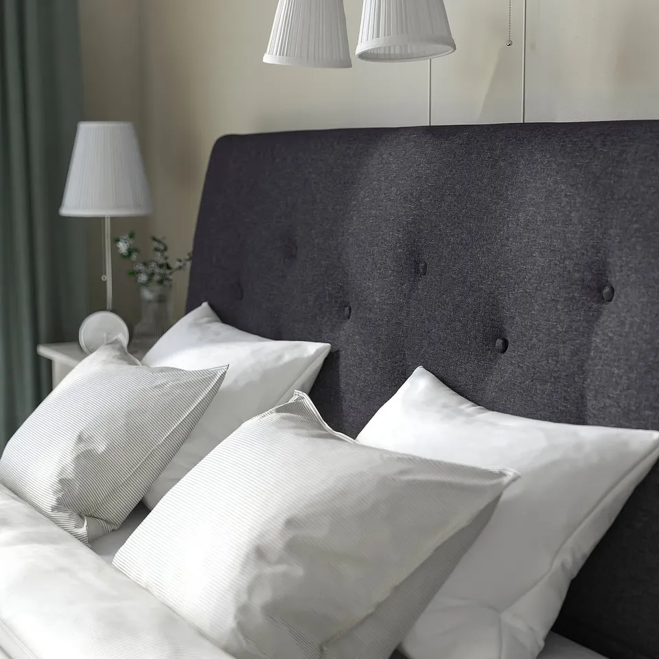 Cosy bed with dark grey headboard and white pillows. Two bedside lamps and a vase with flowers on a small table.