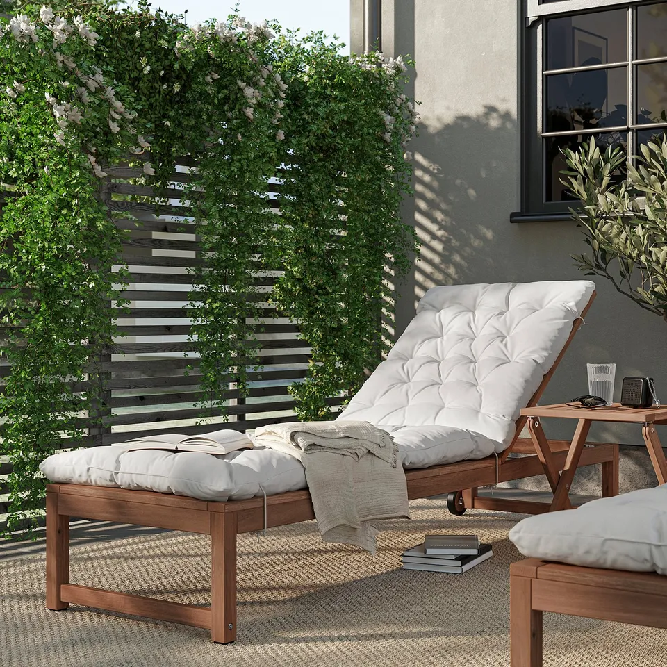 Relaxed outdoor space. Brown wooden recliner and side table, white cushions, greenery background.