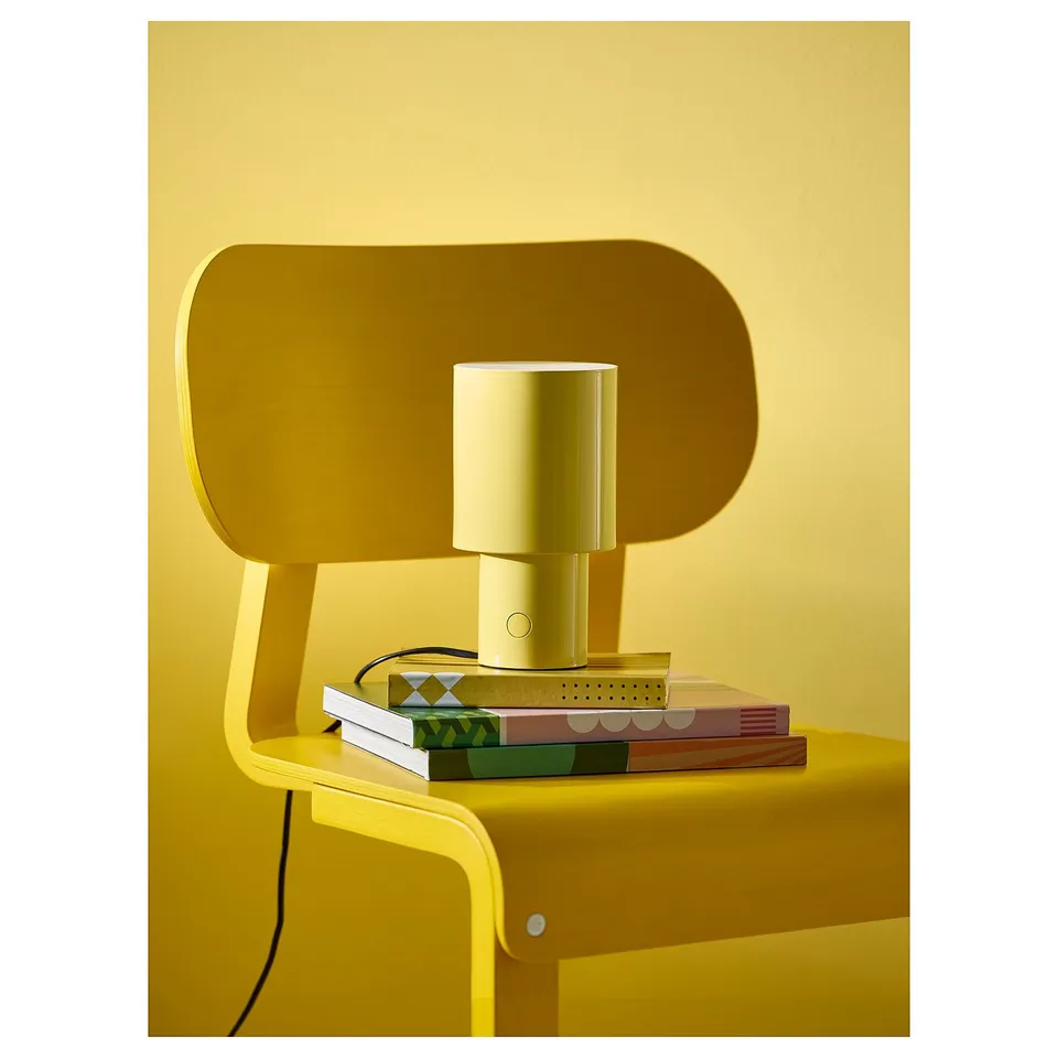 Yellow SPETSBOJ lamp on shelf, emitting soft light, with books and side switch.