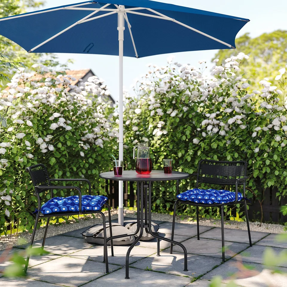Blue patio umbrella with black wicker chairs and floral cushions, set on stone pavers amid white flowering bushes.