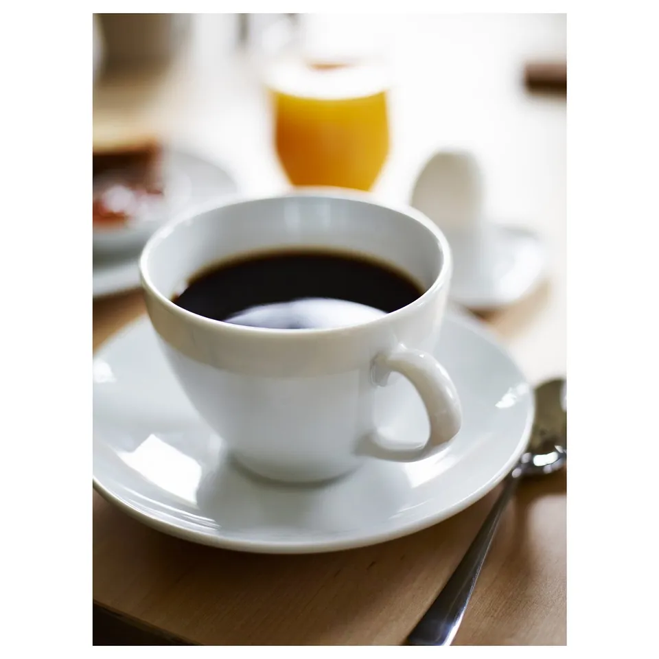 A white porcelain coffee cup on a saucer, made of durable feldspar material. A cutlery set lays beside it on a wooden table.