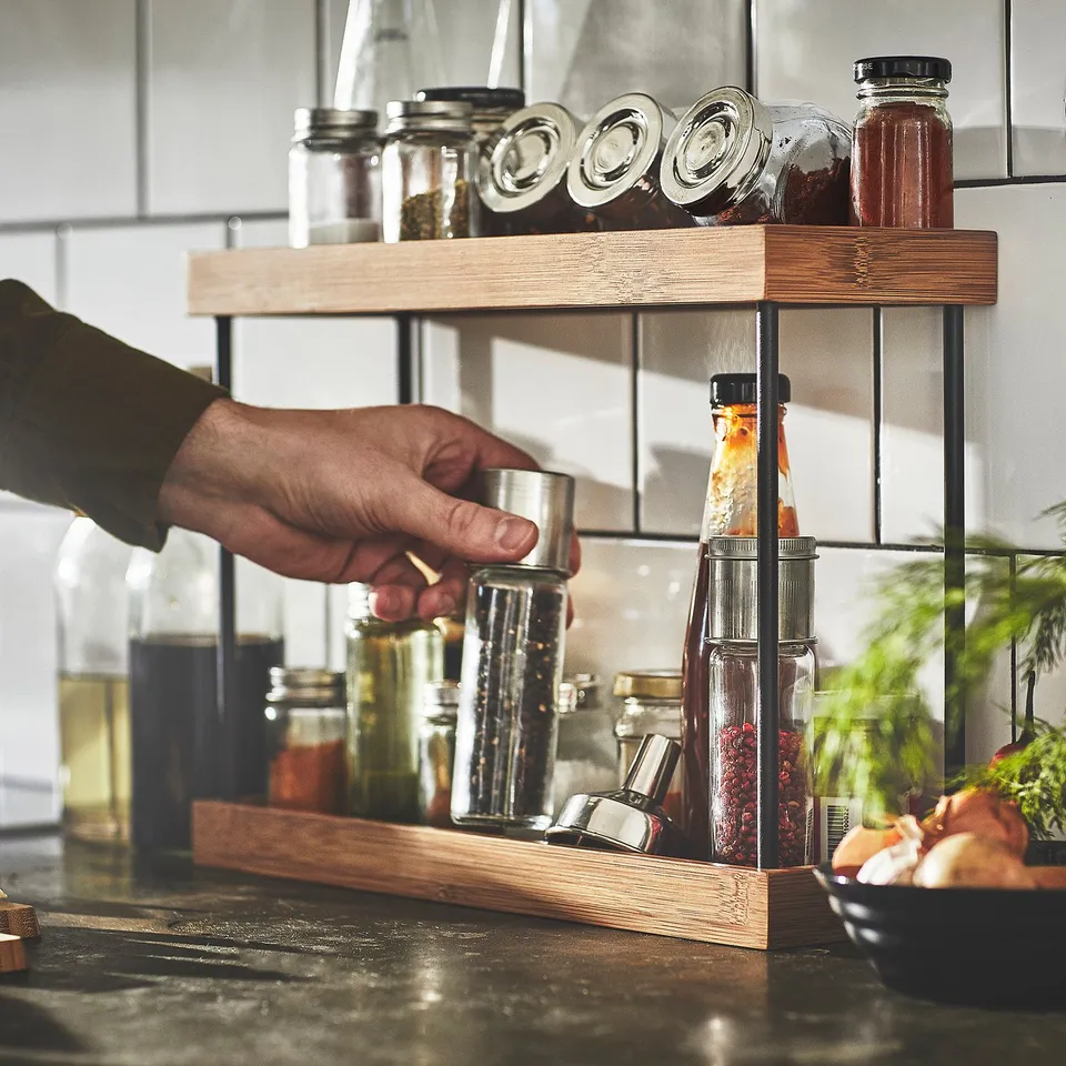 Hand reaching for jar on NÅLBLECKA kitchen organiser with multiple tiers for spice jars and bottles.
