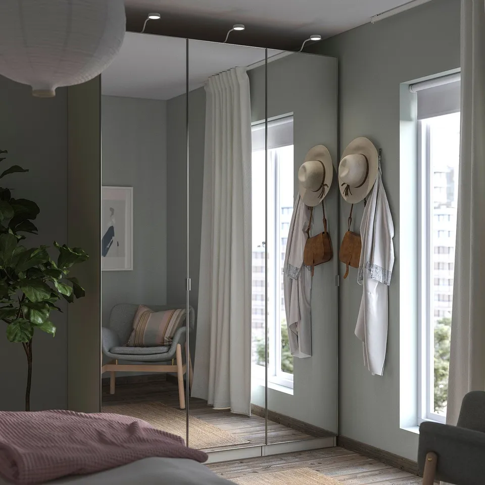 Modern bedroom with grey PAX wardrobe, mirrored doors, hanging hats and bags, plush bed, armchair, and plant.