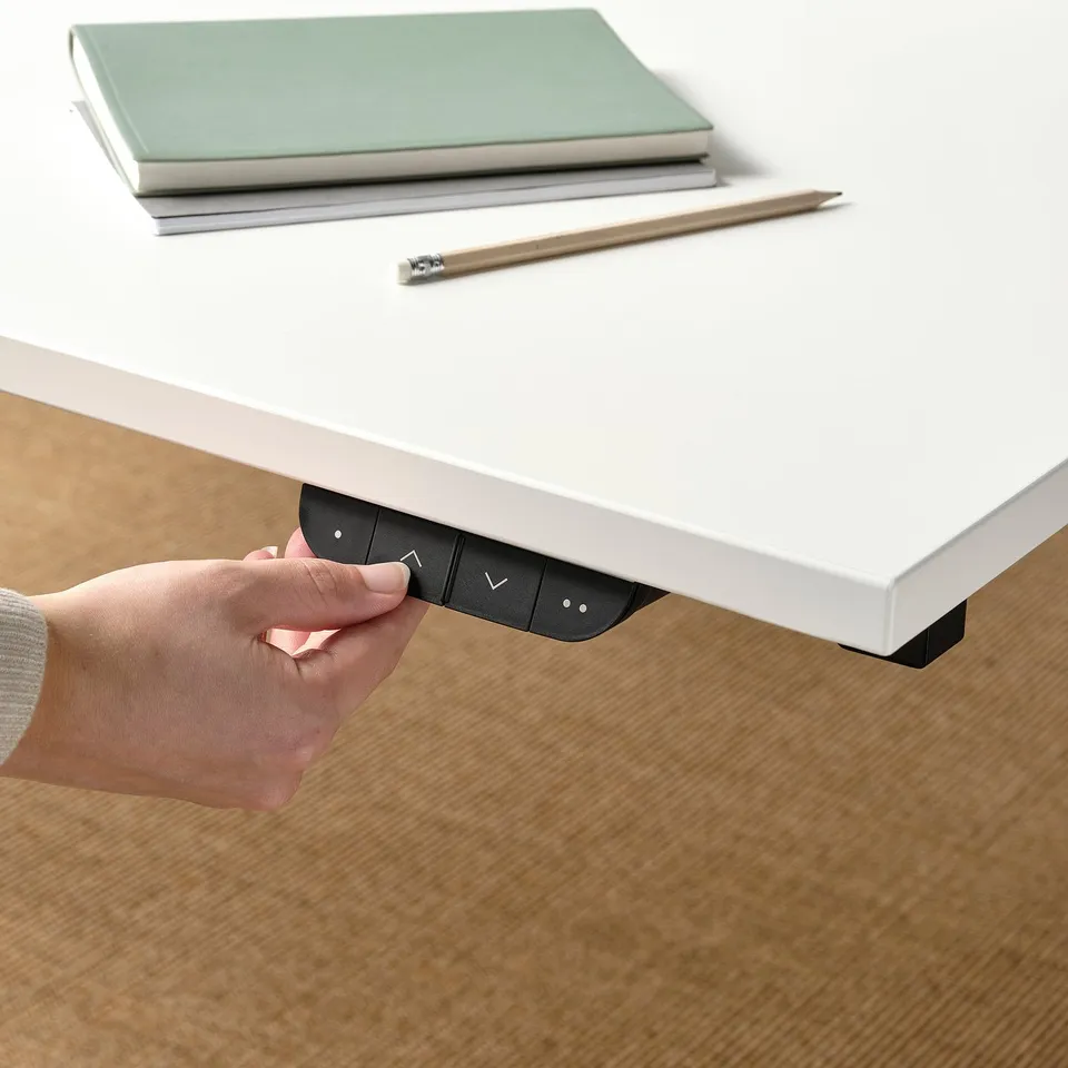 Hand holding black control panel under white desk with texts and symbols, beside closed laptop and pen.