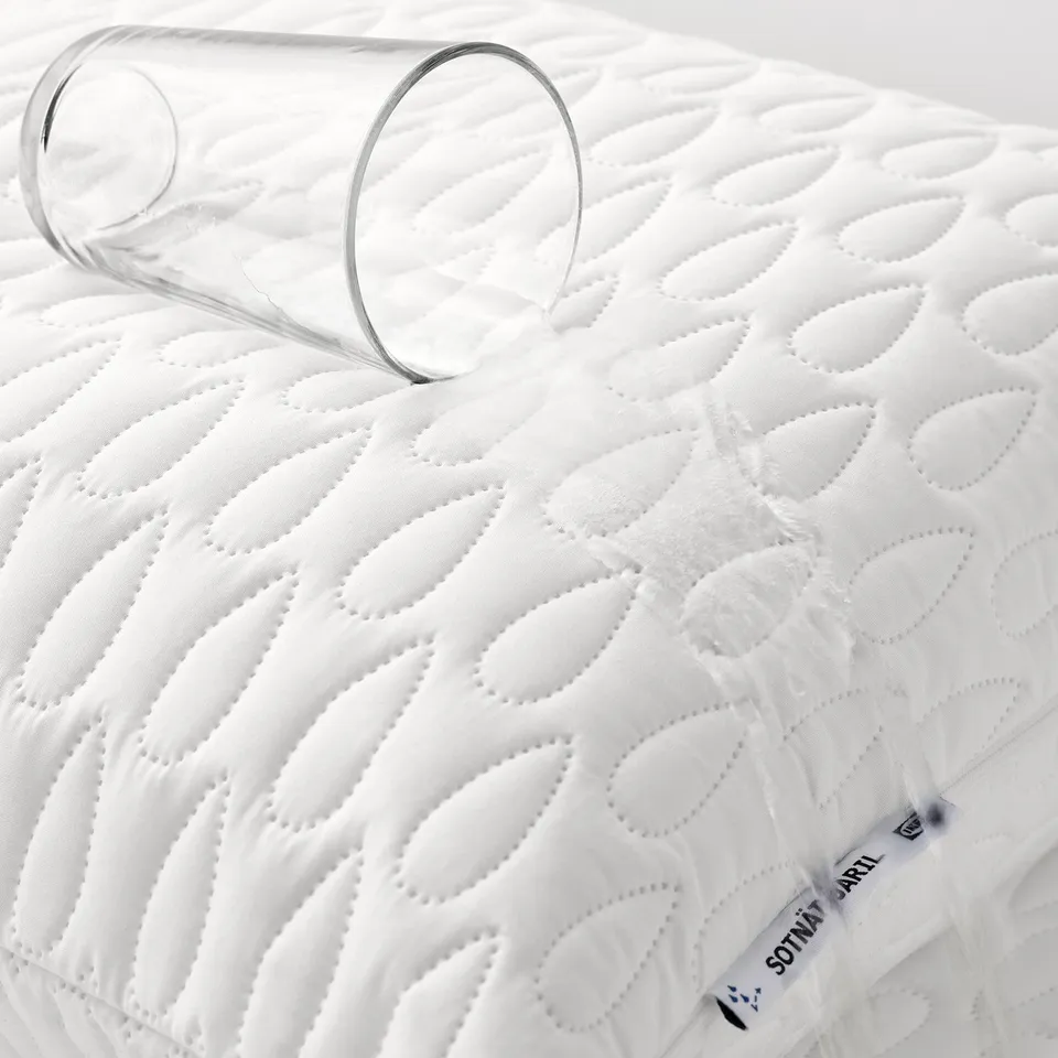 A clear glass is placed on a white quilted pillow protector. The protector has a waterproof layer inside and soft wadding outside.