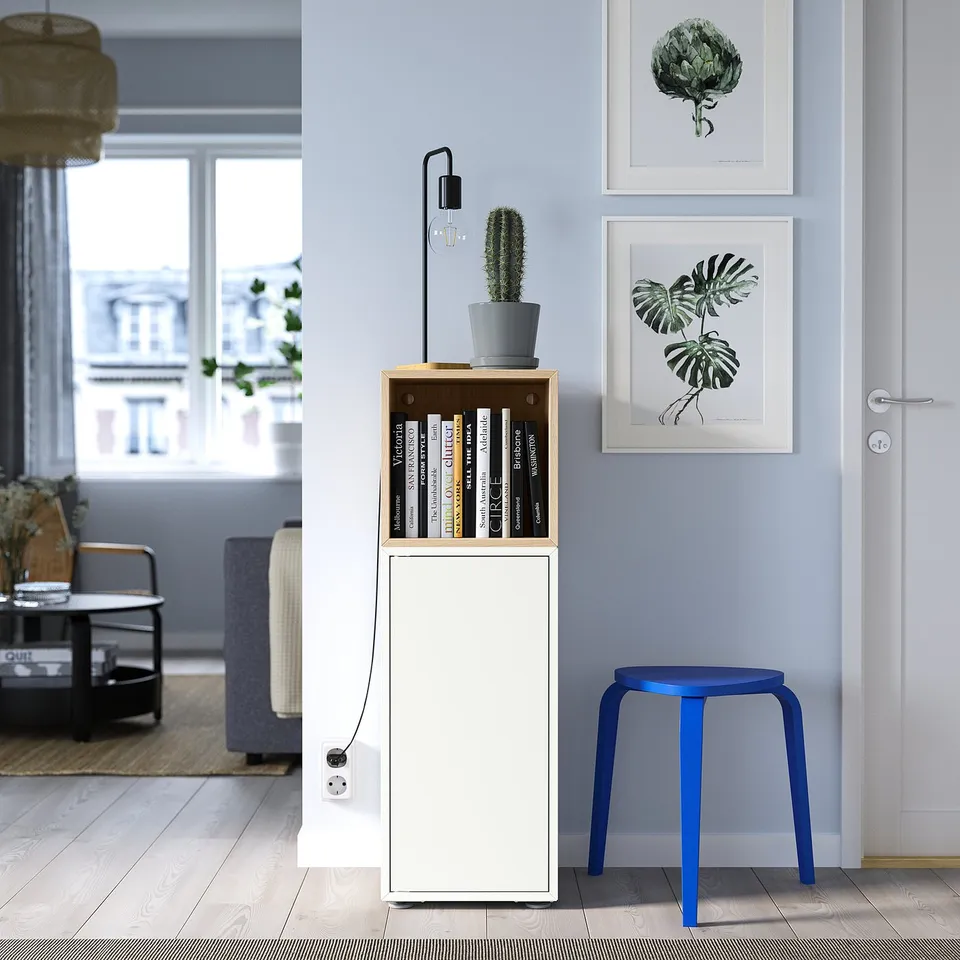 Modern living room with EKET storage, colourful books, blue stool, plant wall art & tall lamp.