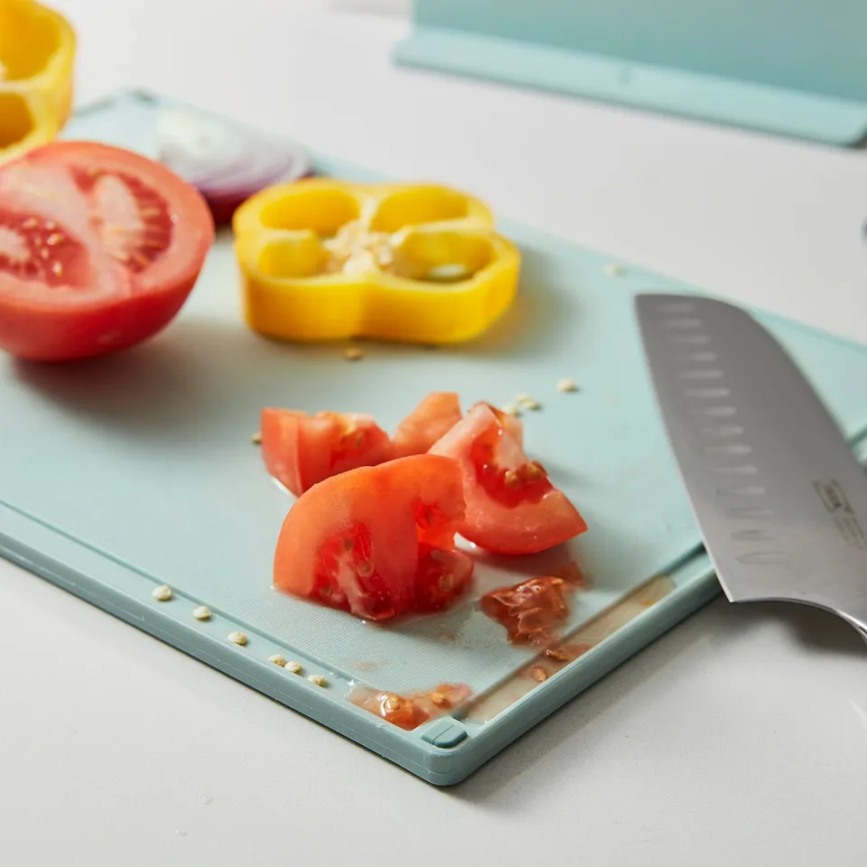 Light blue chopping board with grooves, holding sliced tomatoes and knife. Prevents spills and slips during use.