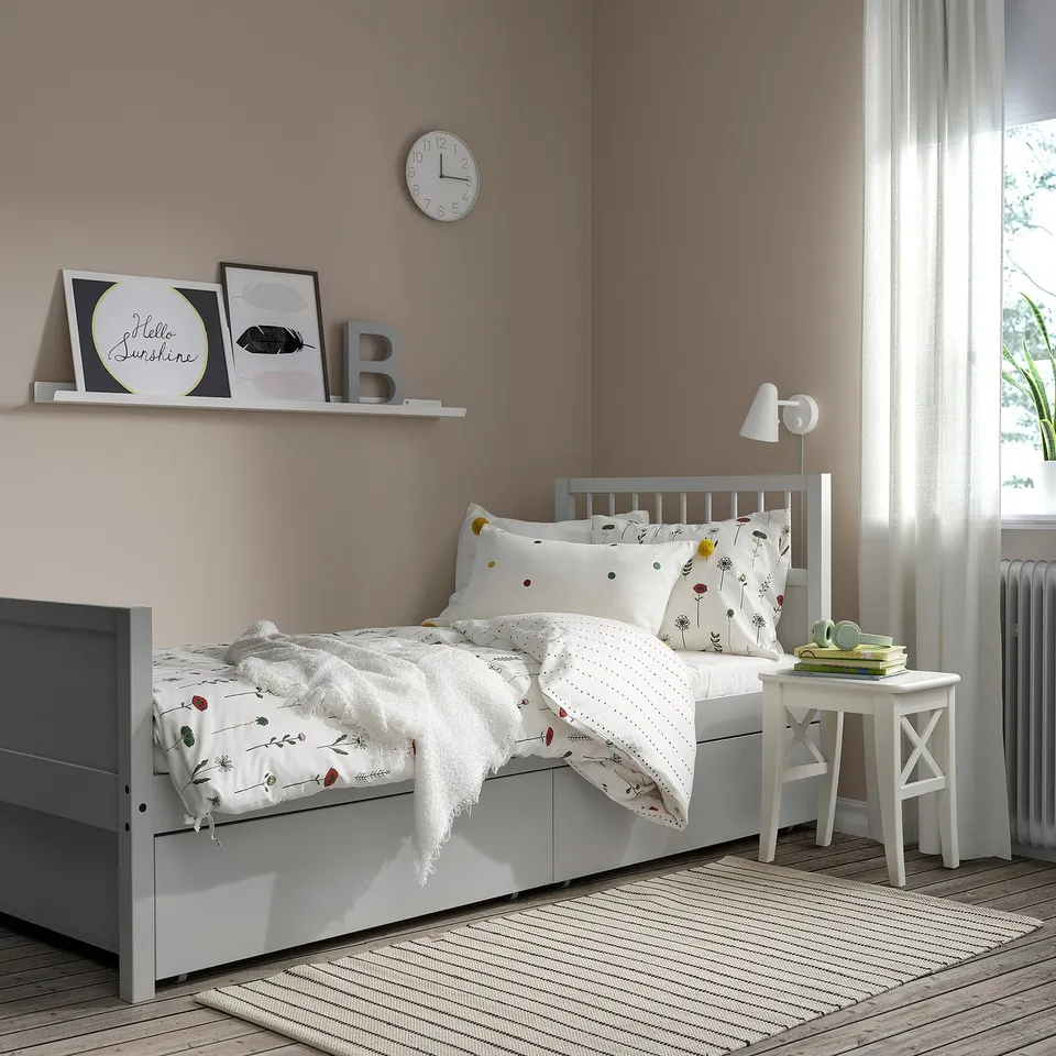 Cosy bedroom with floral bedding, three framed pictures, a clock, striped rug, and a bedside table with a lamp.
